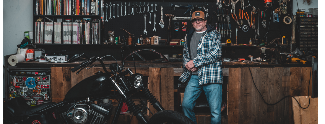 Steve Gerow, artist and co-founder of The Creative Company, standing in his workshop surrounded by an array of tools, a motorcycle, and shelves filled with books and boxes